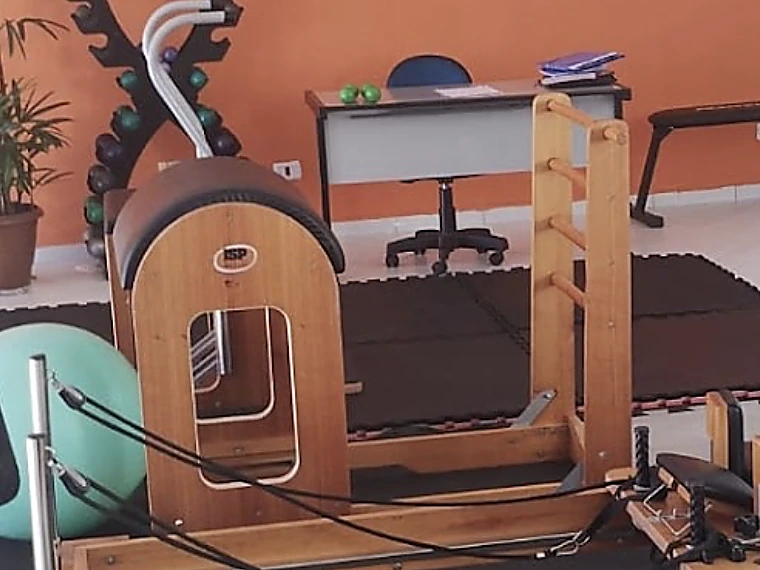 Aparelho de pilates em madeira com escada e apoio curvo, ao lado do rack de halteres