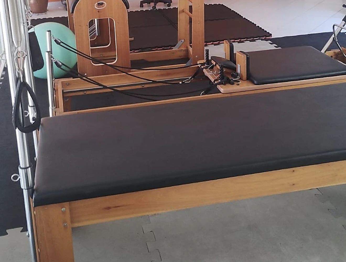 Reformer de pilates com molas e cordas montado na sala, com os demais aparelhos ao fundo